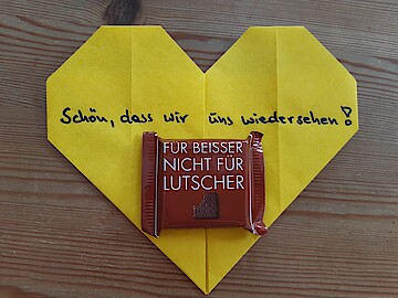 Herz aus farbigem Papier, Aufschrift "Schön, dass wir uns wiedersehen.", eine kleine Schokoladentafel aufgeklebt.