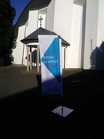 Hintergrund: weiße Kirchenfront, überdachter Eingang zur Kirche; vorne Schatten, aus dem Schatten ragt die "Kirche ist offen"Fahne.