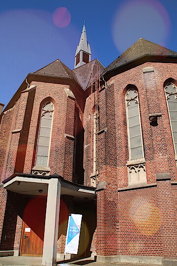 Blick von unten hoch zu der kleinen spitzen Kirchturmhaube, 3 schmale neugotische Fenster, rechts neben dem Eingang steht die Fahne "Kirche ist offen", massive Holztür