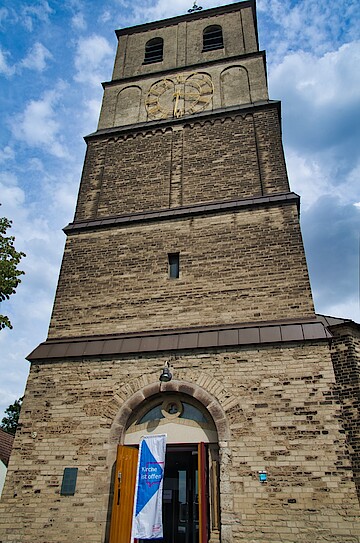 Wuchtiger steinerner Kirchturm eckig, in 4 Etagen, blauer Himmel; offene Kirchentür, davor die Fahne "Kirche ist offen".