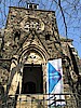 Blick von unten auf ein Kirchenportal, der Turm ragt in einen wolkenlosen Himmel. Rechts neben der hohen Eingangstür steht eine Beachflag "Kirche ist offen"